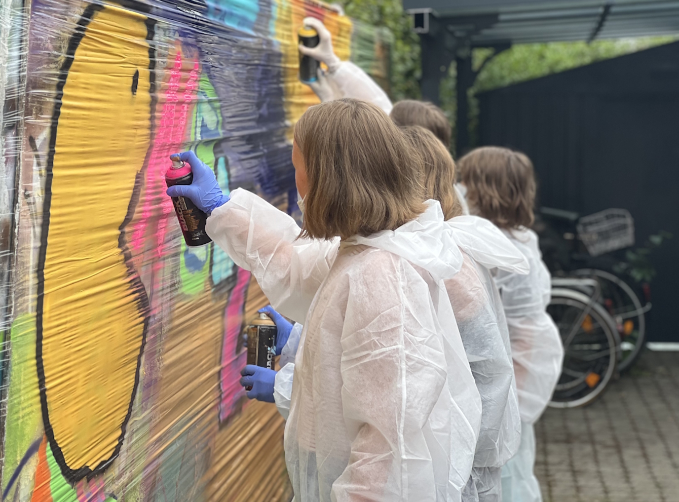 Kinder sprühen an einer Graffitiwand durch weiße Anzüge geschützt