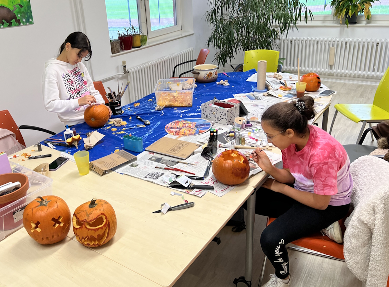 Zwei Mädchen sitzen an einem großen Tisch mit viel Material und gestalten Kürbisfratzen zu Halloween.