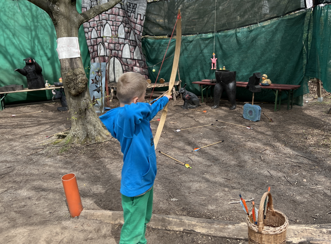 Junge in blauer Jacke und grüner Hose hält einen Pfleil und Bogen in der Hand und zielt auf eine Zielscheibe.