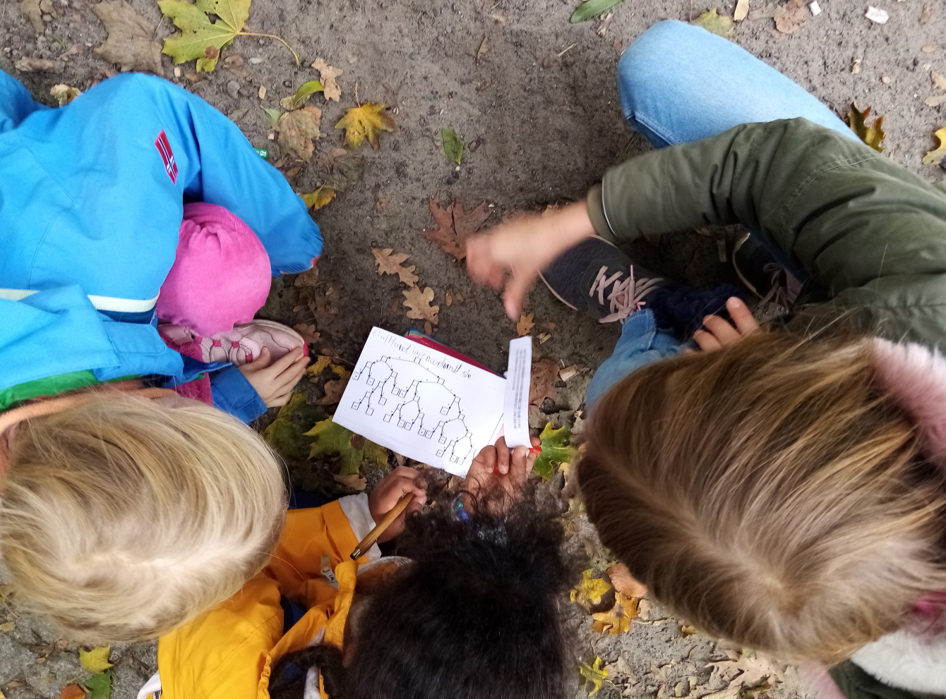 Kindern hocken auf dem Waldboden und schauen auf einen Zettel mit seltsamen Zeichen.