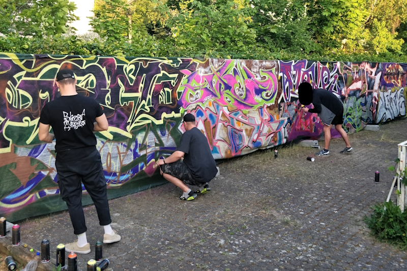 Drei Sprayer sprühen Graffities an eine lange Wand, die auf einem Privatgrundstück steht.