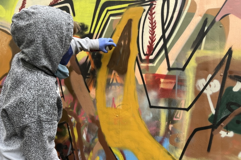 Junge steht seitlich vor einer Graffiti-Wand und sprüht Farbe darauf.