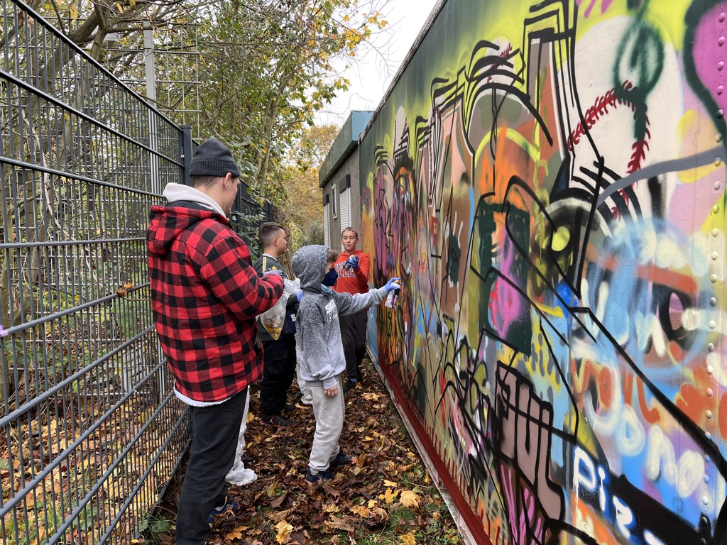 Eine Gruppe von Jungen steht vor eine Graffiti-Wand und besprüht sie. Der Streetart-Künstler neben ihnen leitet an.